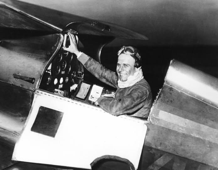 A smiling pilot in goggles and a scarf reaches up to the canopy from an open cockpit