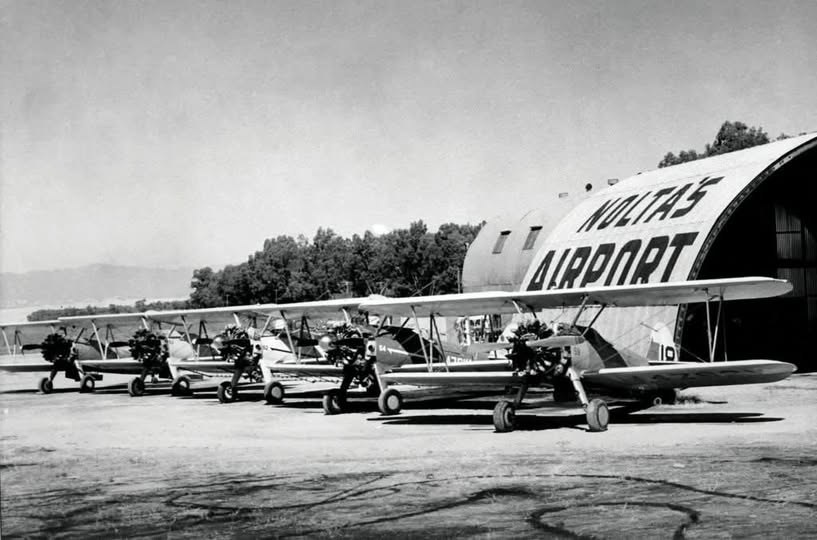 A row of biplanes parked in front of a hangar painted Noltas Airport