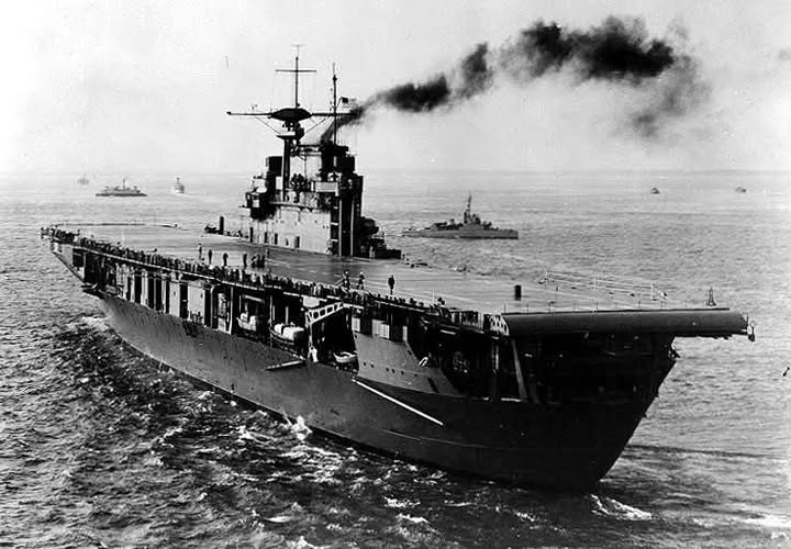 The aircraft carrier USS Hornet underway with smoke trailing from her stack and escort ships behind