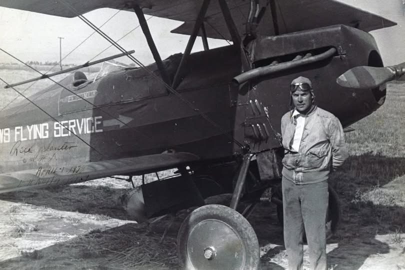 A pilot in goggles and a flight jacket stands beside a Willows Flying Service biplane marked Rice planter
